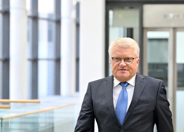 Ein Mann mit Brille und Anzug mit blauer Krawatte steht in einem hellen Gebäude.