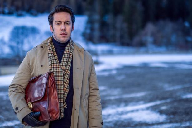 Ein Mann mit Mantel, kariertem Schal und brauner Ledertasche steht draußen vor einer winterlichen Landschaft.