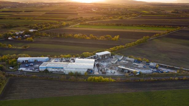 Industrieanlage mit mehreren Hallen und Lagerflächen, umgeben von Feldern und Bäumen bei Sonnenuntergang.