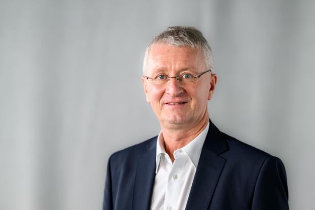 Ein älterer Mann mit Brille, grauen Haaren und Anzug lächelt vor einem hellgrauen Hintergrund.