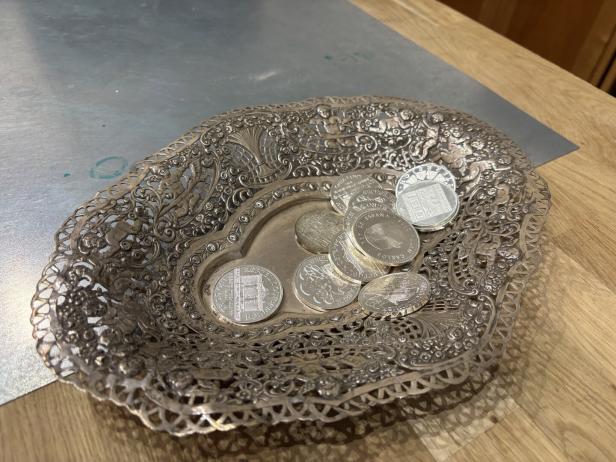 Eine verzierte Silberschale mit mehreren glänzenden Münzen liegt auf einer Holzoberfläche.