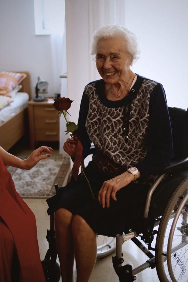 Ältere Frau im Rollstuhl hält eine rote Rose und lächelt, während sie in einem hellen Zimmer sitzt.