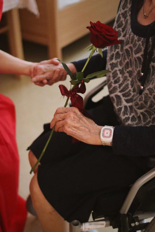 Eine ältere Person im Rollstuhl hält eine rote Rose und wird von einer anderen Person an der Hand gehalten.