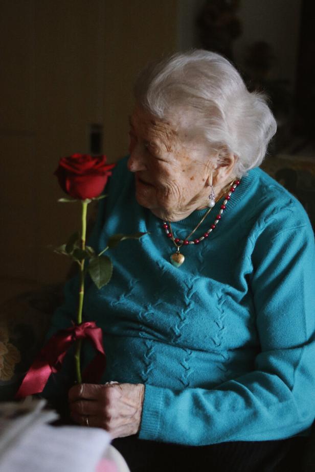 Ältere Frau mit weißem Haar hält eine rote Rose mit Schleife und lächelt sanft.