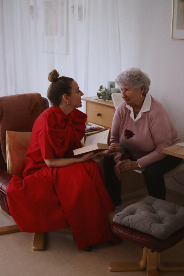 Eine jüngere Frau im roten Kleid liest einer älteren Frau mit grauen Haaren, die eine Rose hält, etwas vor.