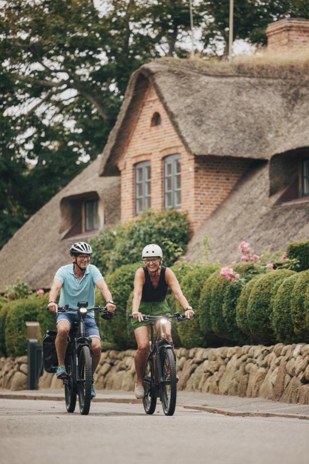 Zwei Menschen fahren mit Fahrrädern und Helmen an einem reetgedeckten Haus mit Garten vorbei.
