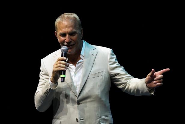 Kevin Costner in hellem Anzug spricht ins Mikrofon und deutet mit der Hand.