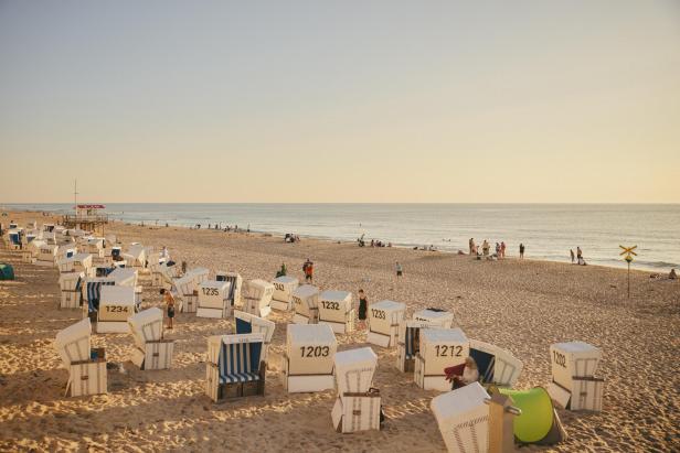 Strandkörbe stehen im Sand an einem ruhigen Strand, während Menschen spazieren gehen und das Meer genießen.