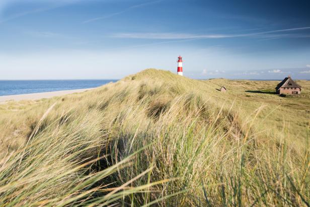 Ein rot-weißer Leuchtturm steht auf einer grasbewachsenen Düne nahe dem Meer, daneben ein reetgedecktes Haus.