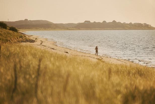 Eine Person joggt am Sandstrand entlang, im Vordergrund hohes Gras, im Hintergrund Hügel mit Häusern.