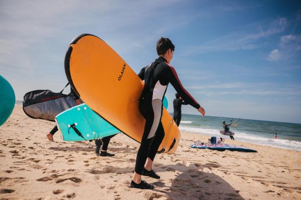 Mehrere Personen in Neoprenanzügen tragen Surfbretter am Sandstrand Richtung Meer, im Hintergrund ein Kitesurfer.