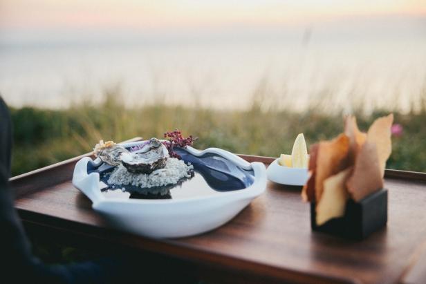 Eine Auster auf grobem Salz mit essbaren Blüten, Zitronenspalten und knusprigen Brotscheiben auf einem Tablett.