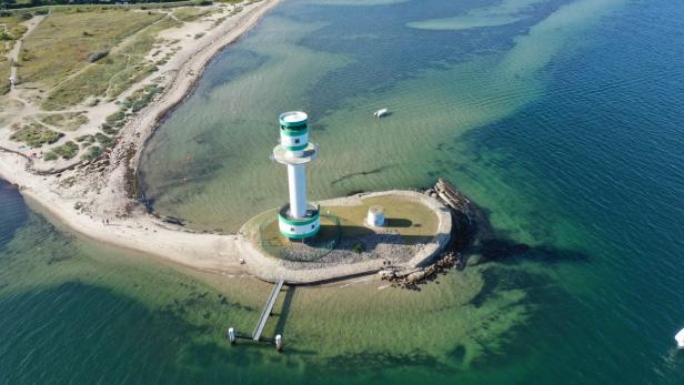 Ein weiß-grüner Leuchtturm steht auf einer kleinen Insel, verbunden mit einem Sandstrand und umgeben von blauem Wasser.
