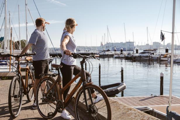 Ein Paar mit Fahrrädern blickt auf einen Hafen mit Segelbooten und einem Schiff.