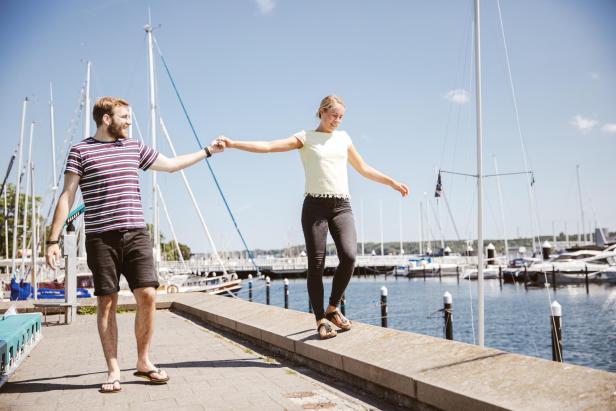 Ein Paar balanciert händchenhaltend auf einer Mauer am Hafen, im Hintergrund Segelboote.