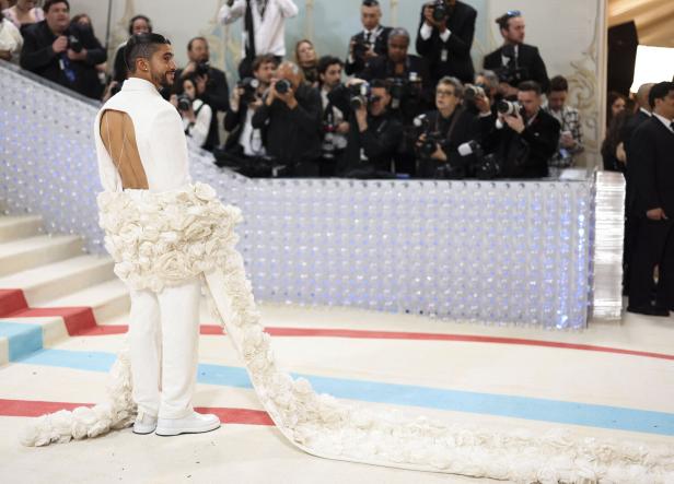 Bad Bunny trägt einen weißen Anzug mit offenem Rücken und langer Schleppe aus weißen Stoffblumen, umgeben von Fotografen.