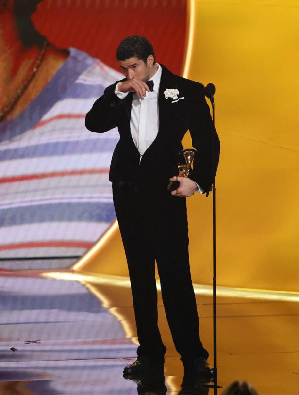 Ein Mann im Smoking - Bad Bunny - steht emotional mit einer Trophäe auf einer Bühne vor einem Mikrofon.