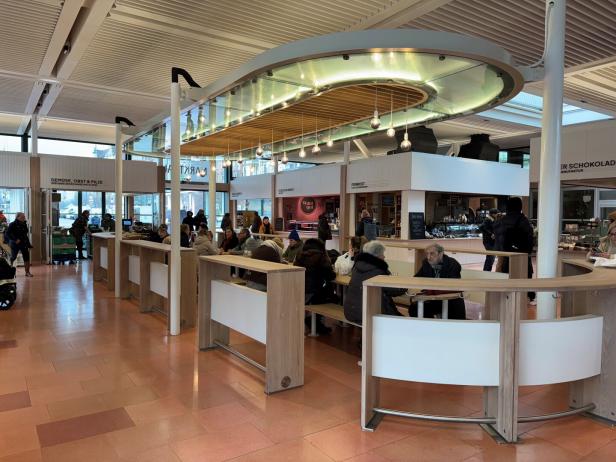 Mehrere Menschen sitzen und essen an Holztischen in einer modernen Markthalle mit verschiedenen Essensständen.