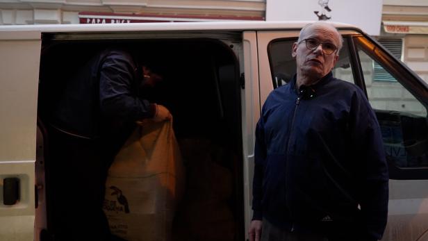 Ein Mann mit Brille steht vor einem Lieferwagen, während eine Person im Inneren einen Sack entlädt.