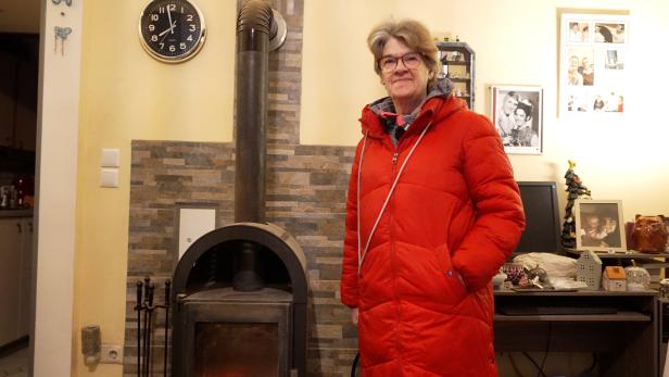 Eine Frau mit roter Jacke steht vor einem Kaminofen in einem Wohnzimmer.