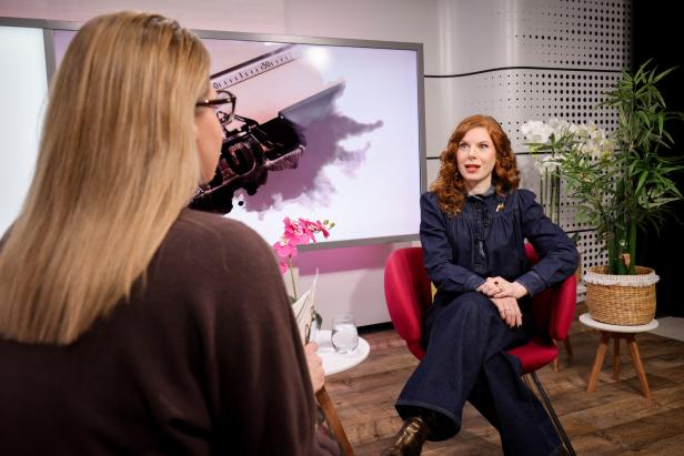Zwei Frauen führen ein Gespräch in einem modern eingerichteten Studio mit Pflanzen und einem großen Bildschirm im Hintergrund.