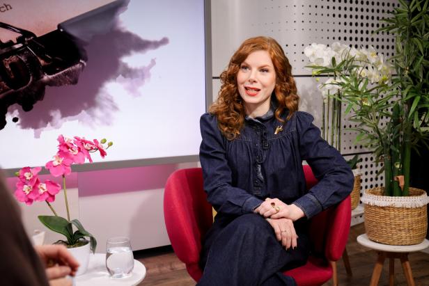 Eine Frau mit roten Locken sitzt in einem roten Sessel, umgeben von Pflanzen und spricht freundlich.