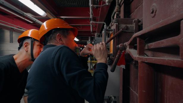 Zwei Arbeiter mit Helmen bedienen eine große, rote Industrieanlage.