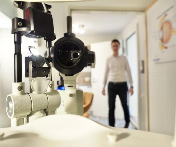 Ein Augenarztgerät steht im Vordergrund, im Hintergrund ist eine Person unscharf zu sehen.