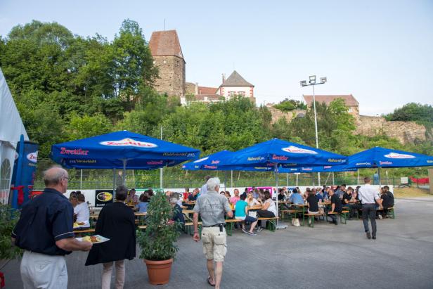 Die Eislaufbahn bei einem früheren Event in den Sommermonaten, Tische sind aufgestellt, Sonnenschirme aufgespannt, Menschen sitzen auf dem Areal verteilt.