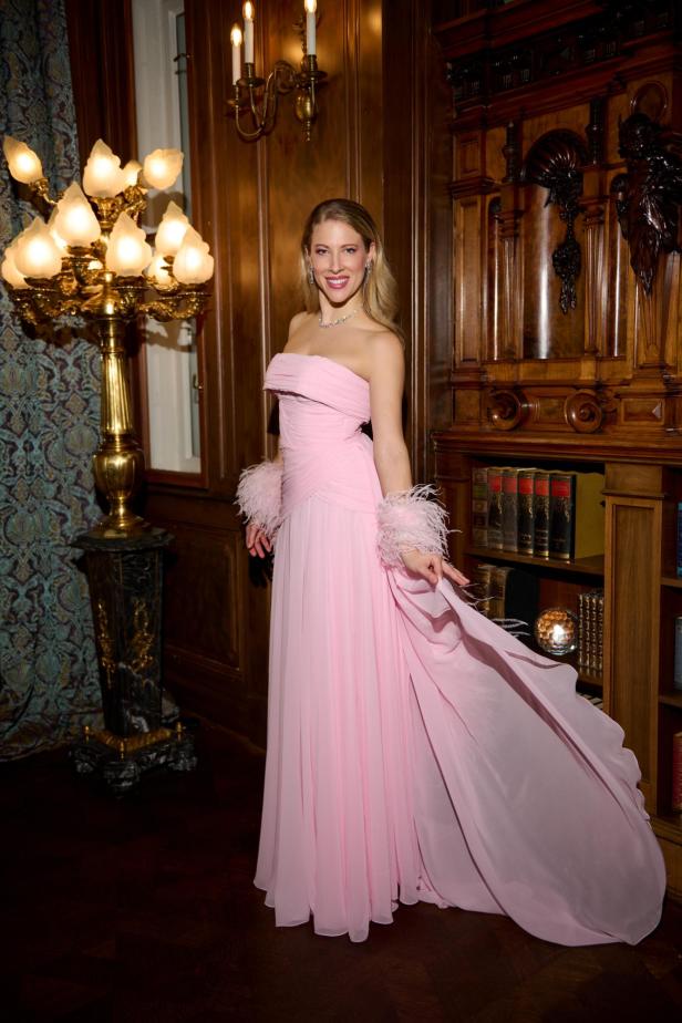 Frau in elegantem rosa Chiffonkleid mit Federboa steht vor holzgetäfelter Wand.