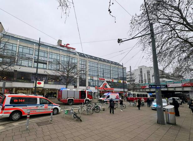 Mehrere Einsatzfahrzeuge von Rettung und Feuerwehr stehen vor einem Einkaufszentrum, während Passanten vorbeigehen.