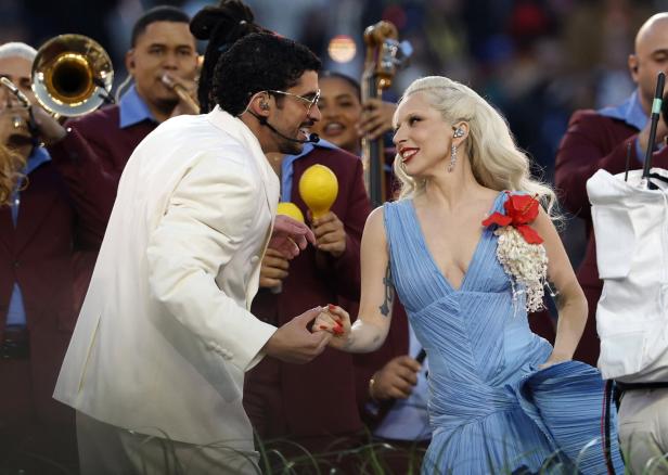 Ein Mann im weißen Anzug und eine Frau im blauen Kleid halten Händchen und lächeln sich auf einer Bühne mit Musikern an.