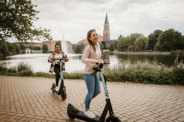 Zwei junge Frauen fahren lachend auf E-Scootern am Kleinen Kiel entlang, im Hintergrund ist der Kieler Rathausturm und ein Springbrunnen zu sehen.