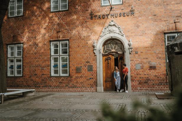 Zwei Personen stehen vor einem historischen Eingang mit Holzportal und unterhalten sich vor einem Gebäude mit der Aufschrift „Stadtmuseum“.