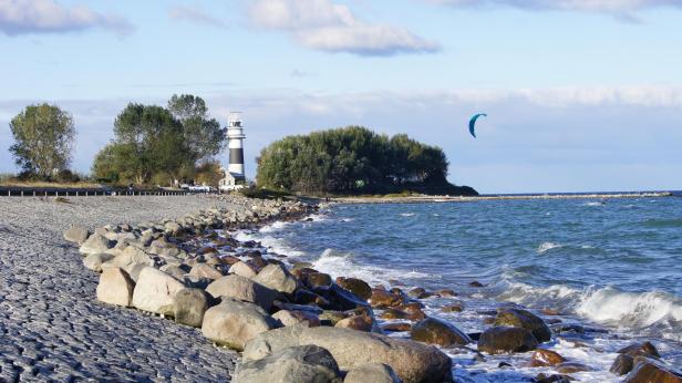 Leuchtturm Bülk an der Ostseeküste mit Wellen, Steinen am Ufer und einem Kitesurfer bei sonnigem Wetter.