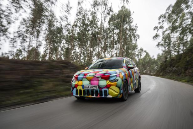 Der Škoda Epiq mit bunter Camouflage-Lackierung fährt auf einer kurvigen Landstraße durch eine bewaldete Landschaft.