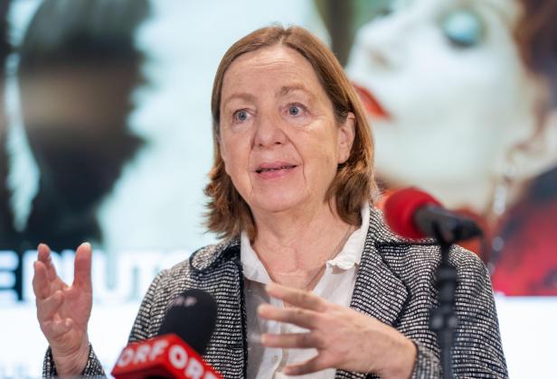 Eine Frau mit schulterlangem Haar spricht gestikulierend vor Mikrofonen, im Hintergrund ist ein unscharfes Porträt zu sehen.