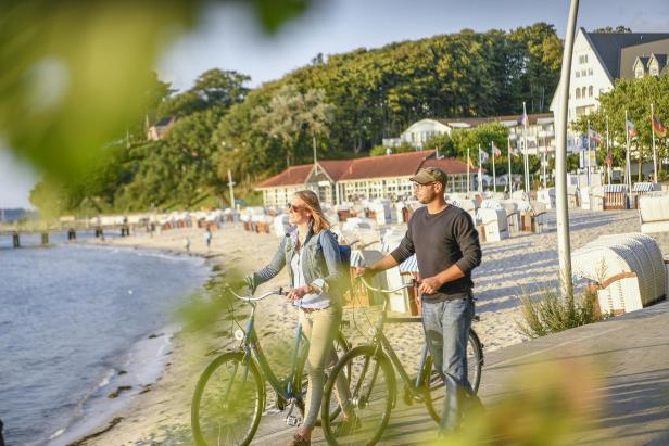 Zwei Menschen schieben ihre Fahrräder an einem Ostseestrand mit Strandkörben entlang, im Hintergrund Bäume und typische Küstenarchitektur.