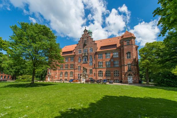 Backsteingebäude mit Giebelfassade und Turm, umgeben von Bäumen und grüner Wiese.