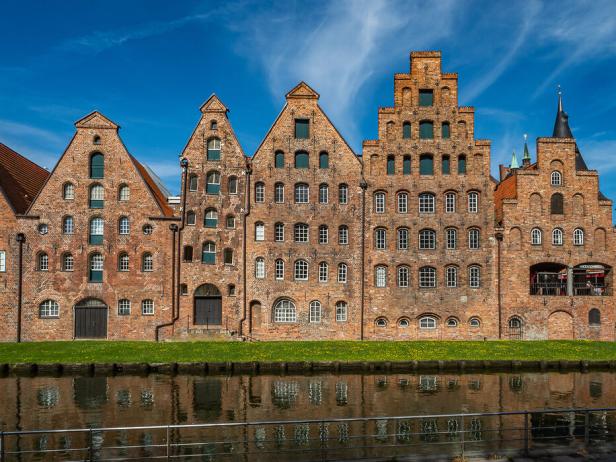 Backsteingebäude mit markanten Giebeln spiegeln sich im Wasser eines Kanals unter blauem Himmel.