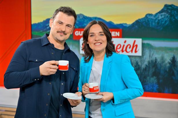 Zwei lächelnde Personen halten Kaffeetassen vor einem Hintergrund mit Bergen und einem Schriftzug.