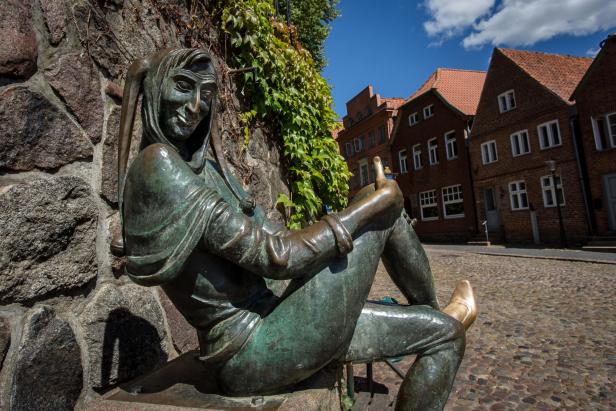 Die bronzene Till-Eulenspiegel-Statue sitzt am Brunnen auf dem Marktplatz von Mölln, umgeben von Kopfsteinpflaster und historischen Backsteinhäusern.