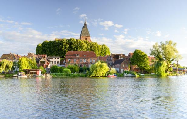Blick auf die Altstadt von Mölln mit historischen Backsteinhäusern, der Kirche St. Nicolai und grüner Uferlandschaft am Wasser.