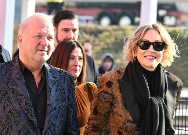 Ein Mann im gemusterten Sakko und eine Frau mit Sonnenbrille und lockigem Haar stehen nebeneinander und lächeln.