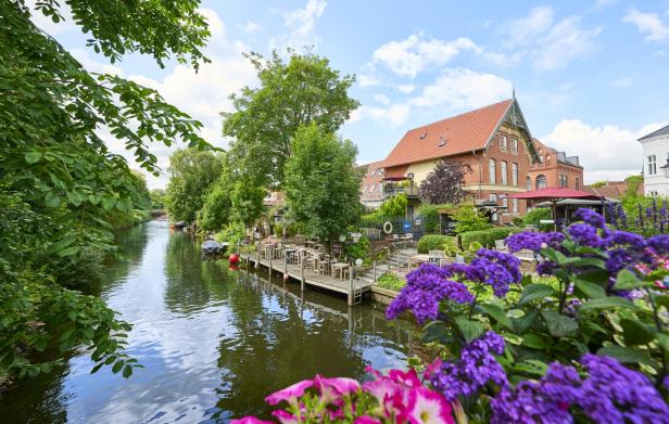 Kanal mit ruhigem Wasser, Holzsteg und Außengastronomie, umgeben von historischen Backsteinhäusern, grünen Bäumen und blühenden Blumen im Vordergrund