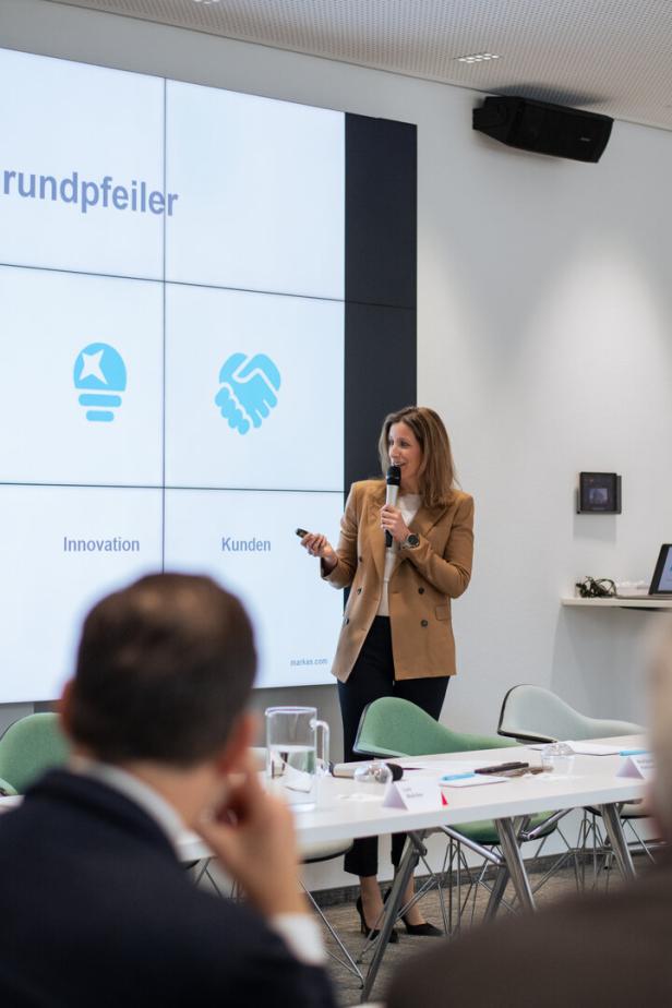Eine Frau hält eine Präsentation vor einem großen Bildschirm mit Symbolen und den Wörtern „Innovation“ und „Kunden“.