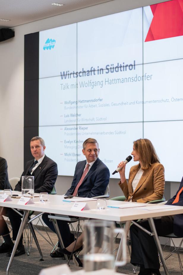 Mehrere Personen sitzen an einem Konferenztisch, während eine Frau mit Mikrofon spricht und im Hintergrund eine Präsentation angezeigt wird.