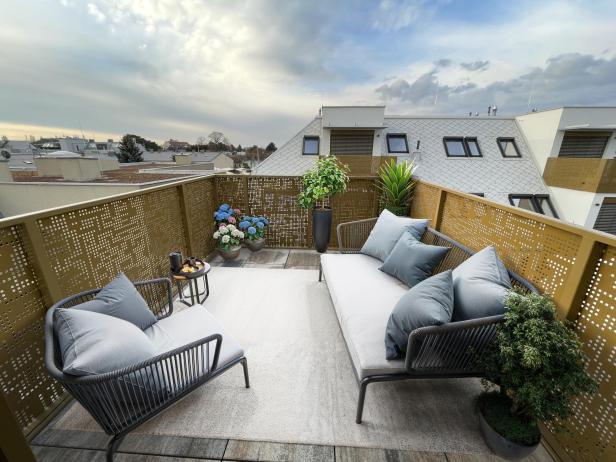 Zwei moderne Sofas mit Kissen, Pflanzen und einem kleinen Tisch mit Getränken auf einem Balkon mit Ausblick auf Dächer.