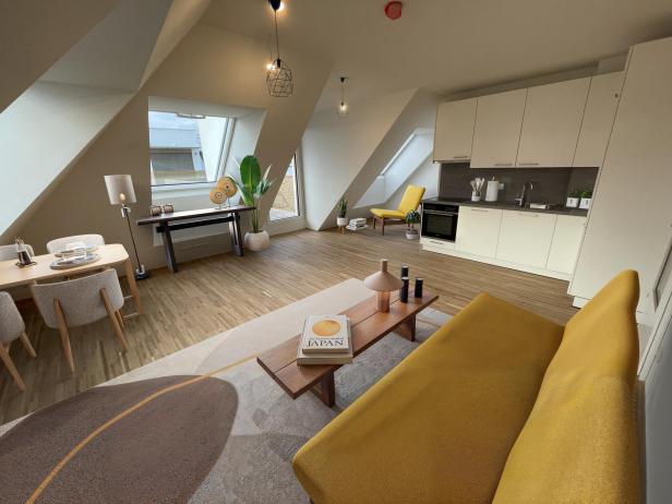 Helles, modern eingerichtetes Wohnzimmer mit gelbem Sofa, Essbereich, offener Küche und großen Fenstern unter Dachschrägen.