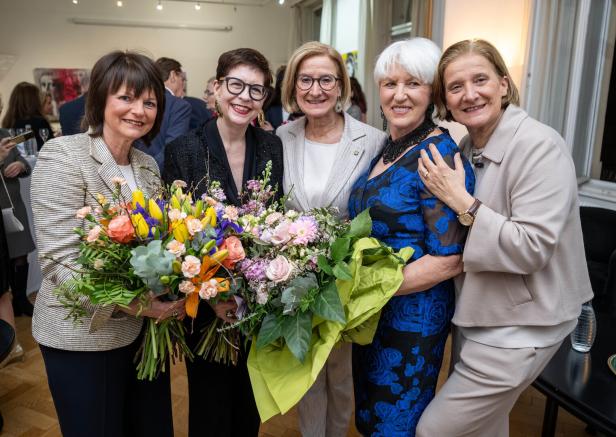 Fünf Frauen stehen eng beisammen, zwei von ihnen halten große bunte Blumensträuße und alle lächeln.
